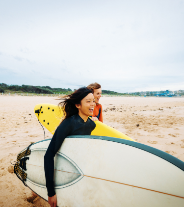 two women walk with their surfboards on a sandy shore, insured with travel insurance for Australia 