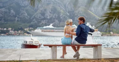 A smiling couple on a bench with a cruise ship in the background, which may have them wondering, "How safe is cruising?".