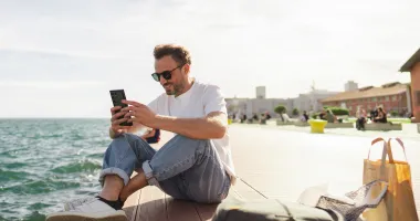 Young man on the phone in front of the ocean searching the difference between a travel alert and a travel advisory