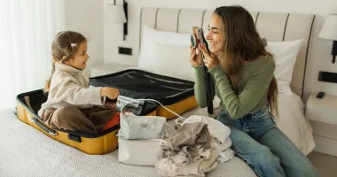 Woman who may have travel anxiety smiling and taking a picture of her young daughter sitting in a suitcase while packing