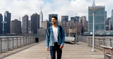 Young man smiling and walking on a bridge in front of the New York City skyline after reading a United States travel guide