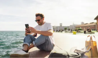 Young man on the phone in front of the ocean searching the difference between a travel alert and a travel advisory