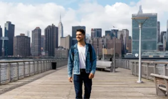 Young man smiling and walking on a bridge in front of the New York City skyline after reading a United States travel guide