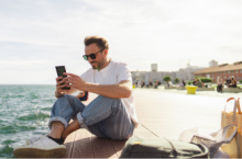 Young man on the phone in front of the ocean searching the difference between a travel alert and a travel advisory