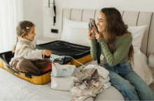 Woman who may have travel anxiety smiling and taking a picture of her young daughter sitting in a suitcase while packing
