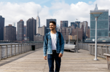 Young man smiling and walking on a bridge in front of the New York City skyline after reading a United States travel guide
