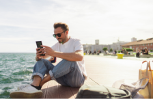 Young man on the phone in front of the ocean searching the difference between a travel alert and a travel advisory