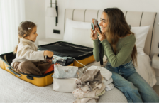 Woman who may have travel anxiety smiling and taking a picture of her young daughter sitting in a suitcase while packing