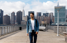 Young man smiling and walking on a bridge in front of the New York City skyline after reading a United States travel guide